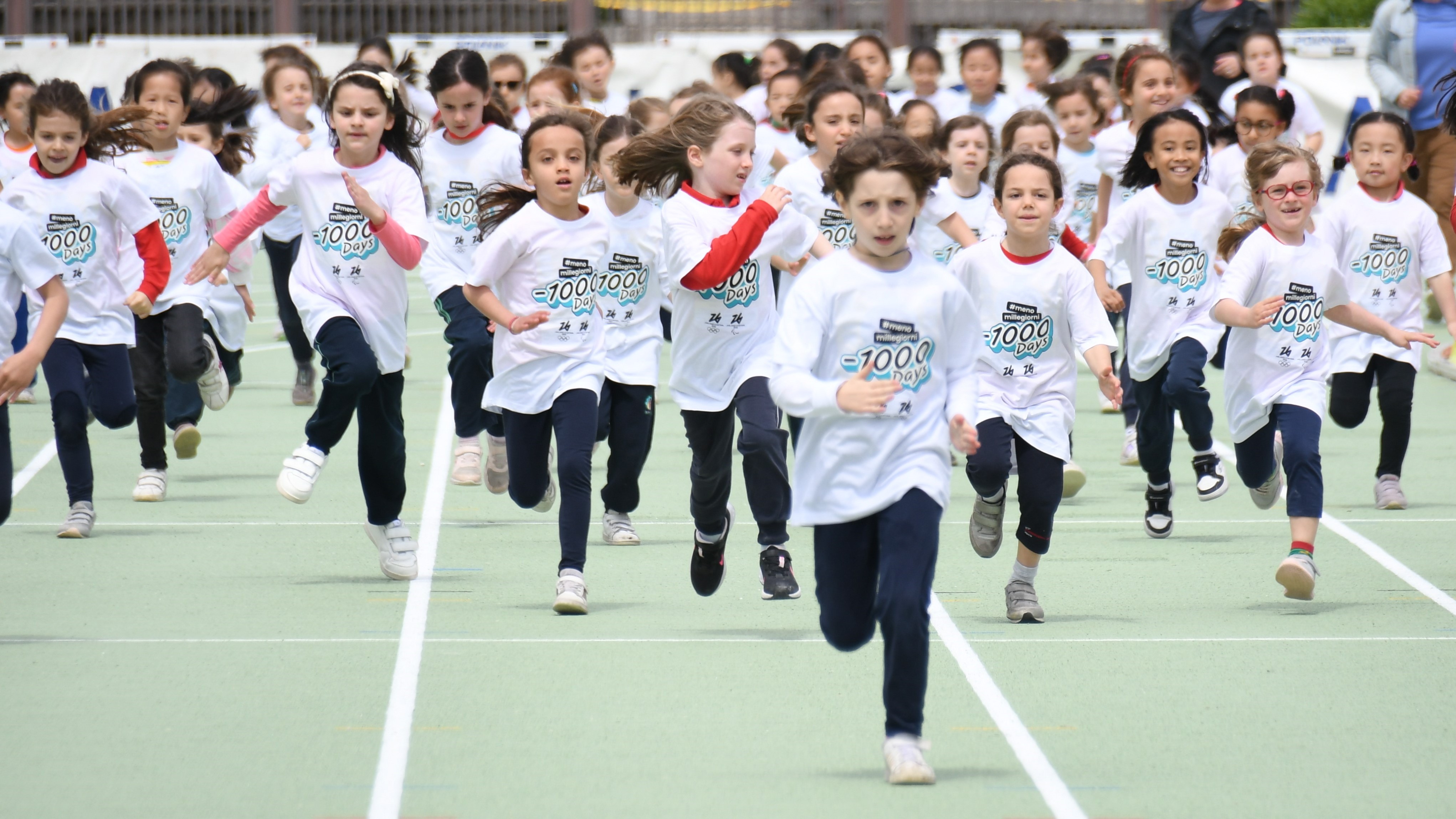 Dei bambini che corrono su un campo di atletica con la maglia di Milano Cortina 2026