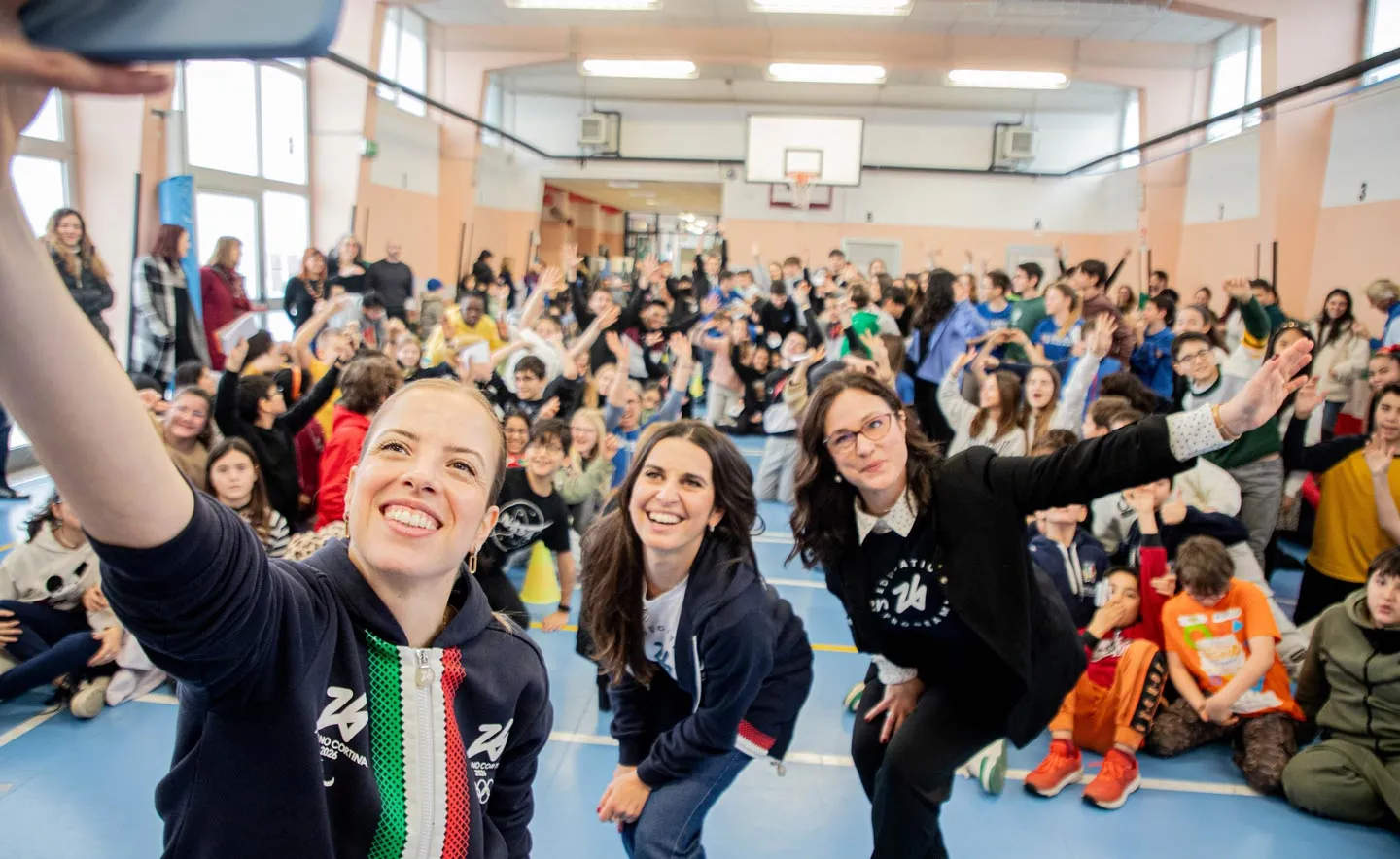 Les travailleurs de Milano Cortina 2026 et Carolina Kostner prennent un selfie avec des enfants dans un gymnase.